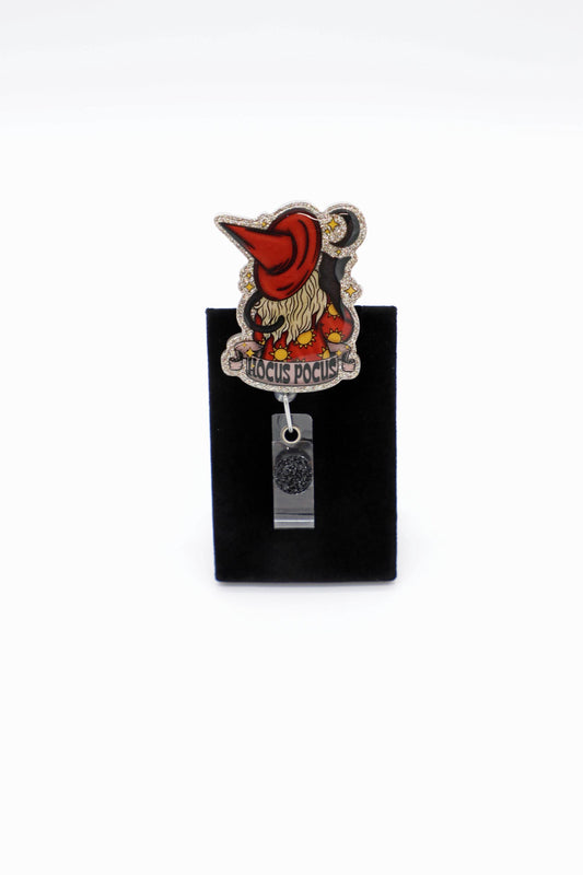 Halloween badge reel magic for your workday. This Hocus Pocus design sparkles with gold  glitter and cozy spooky charm. A crimson witch hat tips over a flowing blonde hair, framed by twinkling stars and a crescent moon. Perfect for anyone who loves Halloween season, a hint of black cat energy, and cute witchy accessories that stand out. Badge reel clip options: Clear Swivel Alligator Clip, Black Swivel Alligator Clip, Clear Belt Clip, White Belt Clip, or White MRI safe clip.